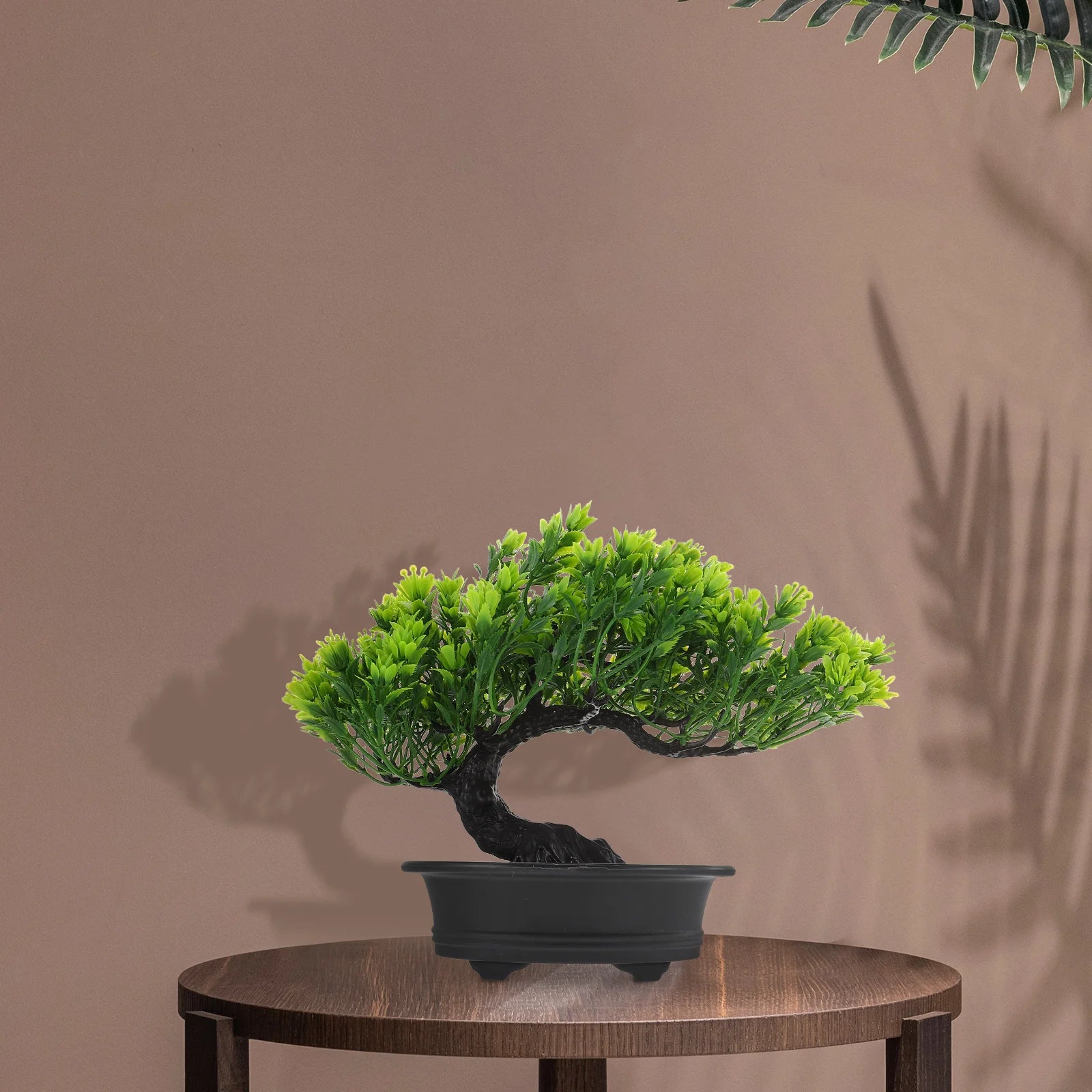 Artificial green bonsai tree in a matte black rectangular pot, set on a wooden table against a soft brown background.