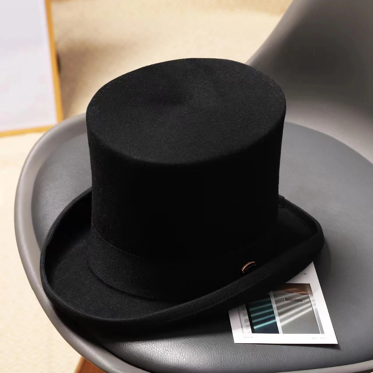 A classic black wool top hat with a flat crown, curved brim, and a matching fabric band, resting on a grey chair.