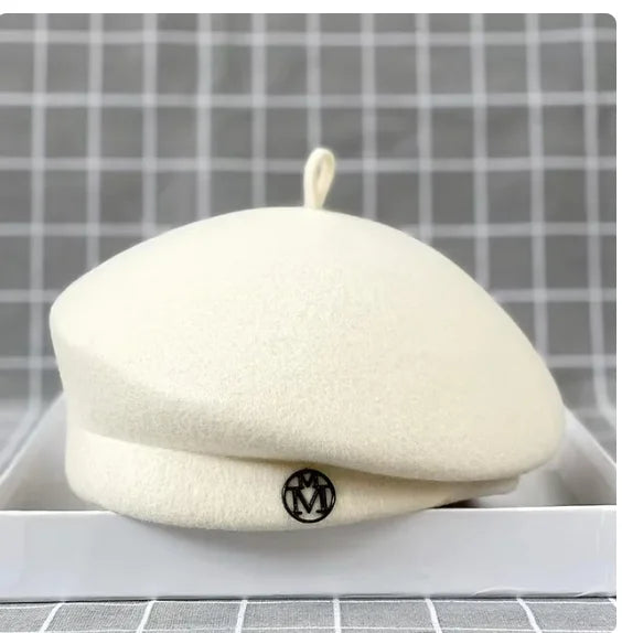 Classic cream-coloured wool felt beret with a small stem detail and a circular 'M' logo badge on the front.