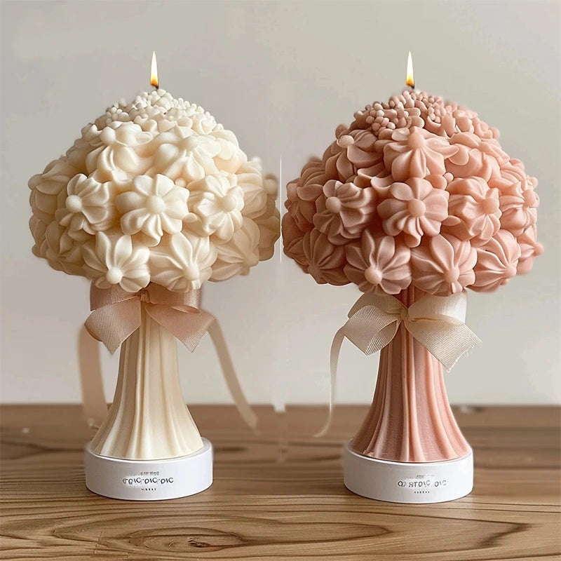 Two 3D floral bouquet pillar candles with ribbon bows, one cream and one pink, standing on wooden surfaces.