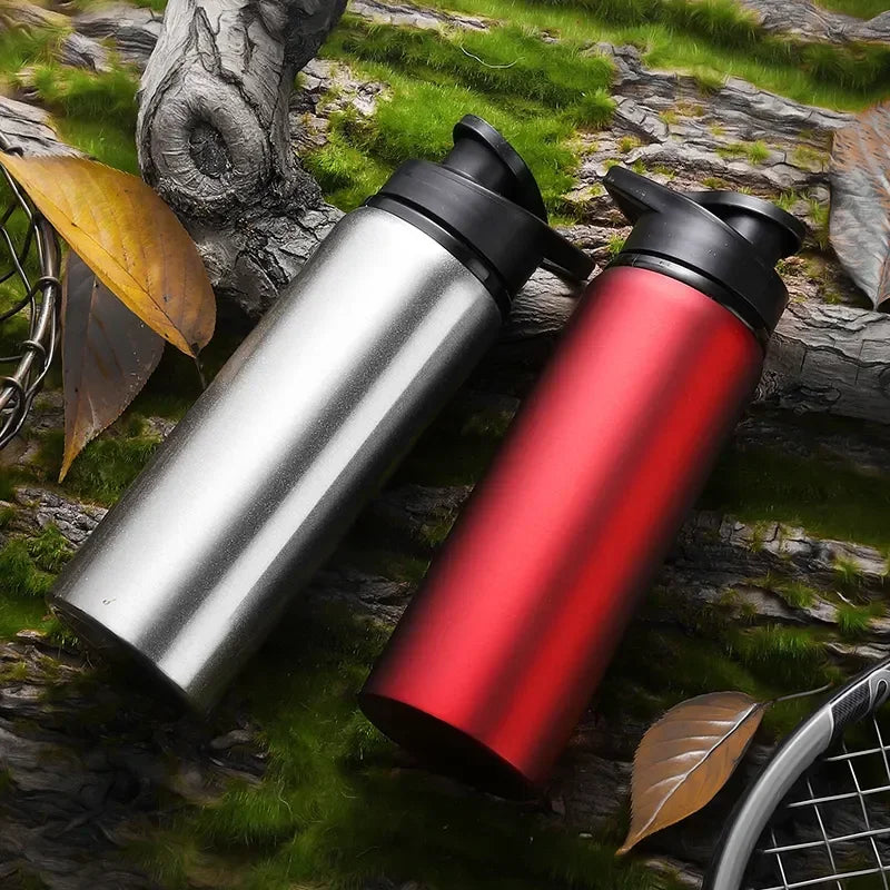 Two metallic sports water bottles, one silver and one red, with black flip-top lids, resting on a mossy log.