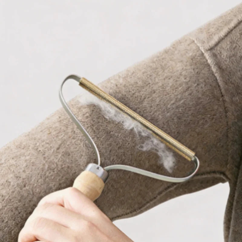 A wooden-handled fabric shaver with a gold metal blade removing lint from a beige wool sleeve.