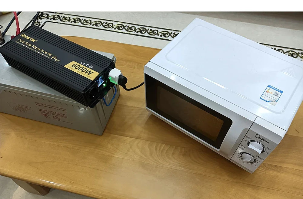 Black 6000W power inverter connected to a battery, powering a white microwave on a wooden table.