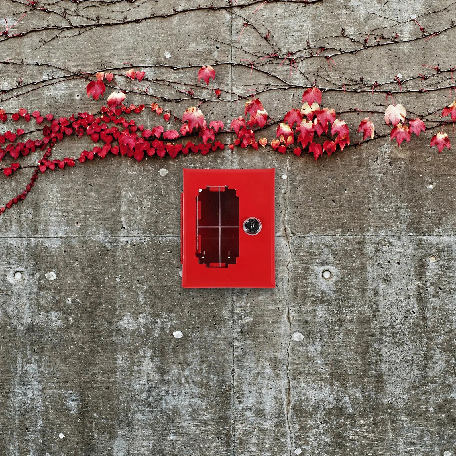 Red metal fire alarm pull station box with a central glass window and key lock, mounted on a concrete wall.