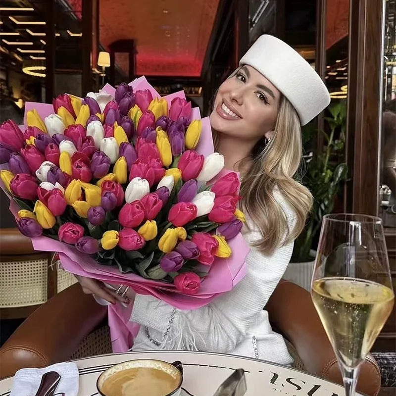 A woman wearing an elegant, structured cream wool hat, holding a large bouquet of multi-coloured tulips.