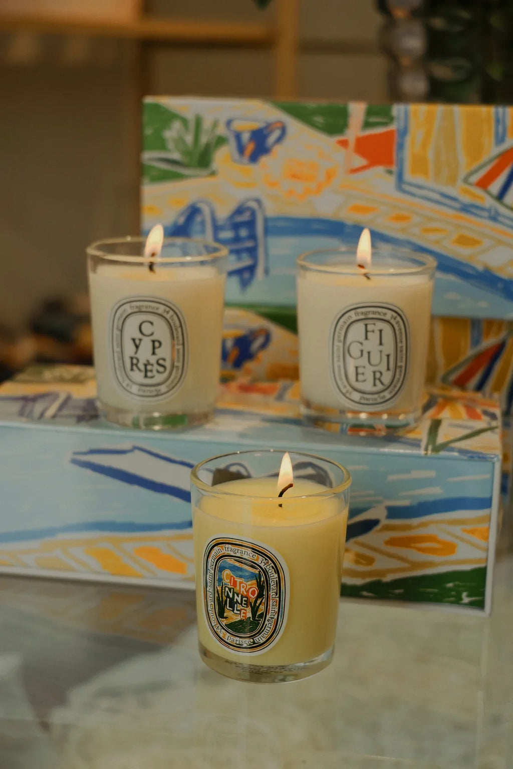 Three scented candles in glass jars with colorful illustrated labels, lit and arranged before a vibrant gift box.