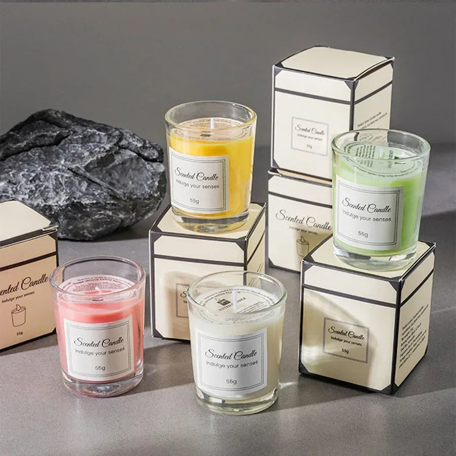 Four colourful scented candles in glass jars with matching gift boxes, set against a grey background.