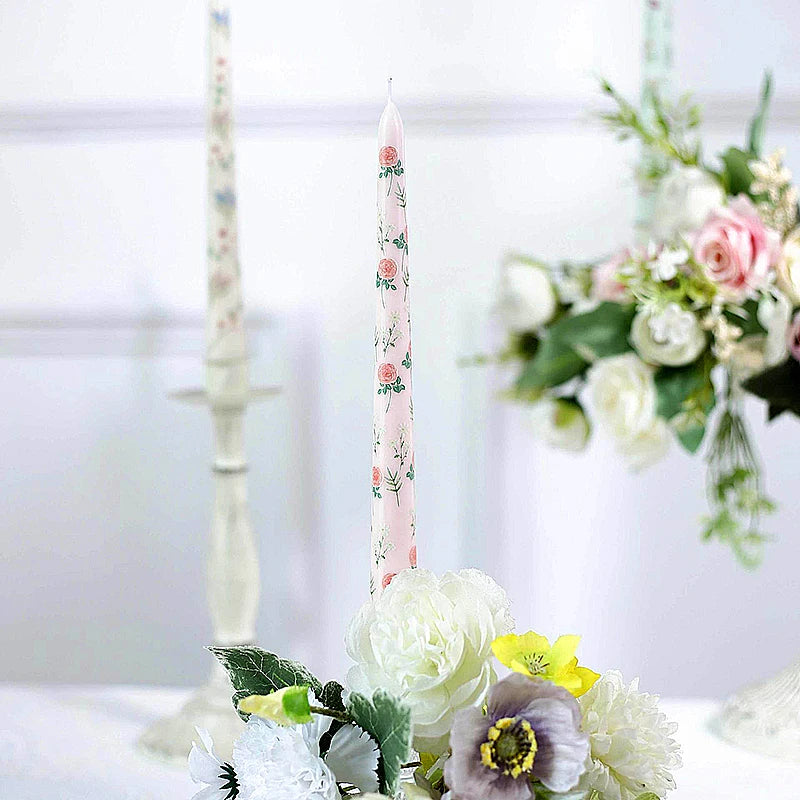 A tall, slim pale pink taper candle featuring a delicate rose flower pattern, styled amidst a floral table arrangement.