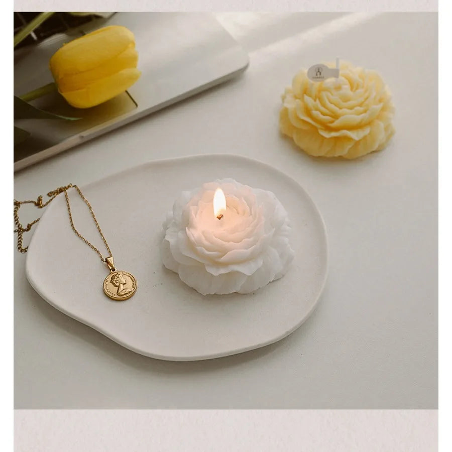 Two elegant rose-shaped candles in white and yellow, displayed on a ceramic tray with a gold coin necklace.