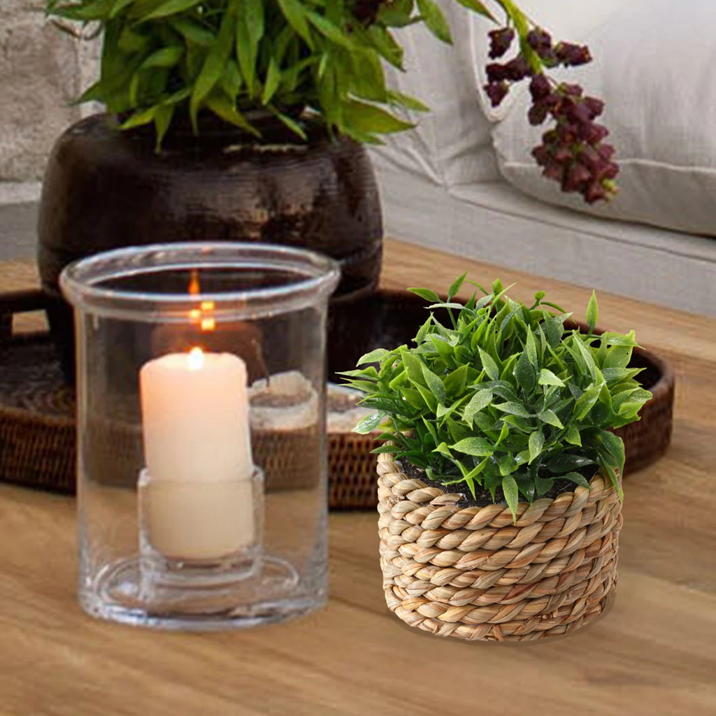 A small, bushy artificial green plant in a rustic woven rope basket, sitting on a wooden table next to a glass candle.