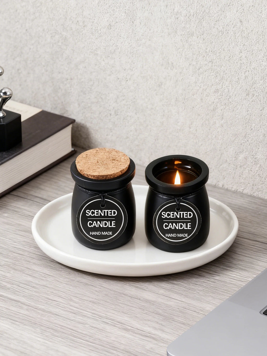 Two black jar candles on a white plate, one lit and one with a cork lid, featuring "Scented Candle" tags.