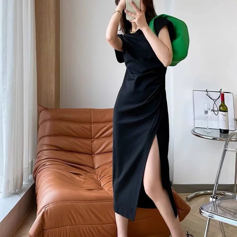 Woman wearing a black midi-length t-shirt dress with a ruched side and a high leg slit, accessorised with a green bag.