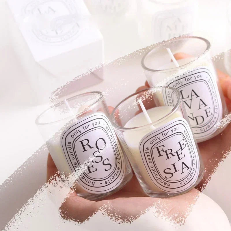 Three clear glass scented candles held in hands, featuring white wax and oval labels with black minimalist text.