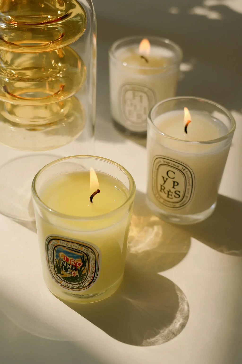 Three lit scented candles in glass jars with oval labels, arranged on a light surface with a golden glass decanter.