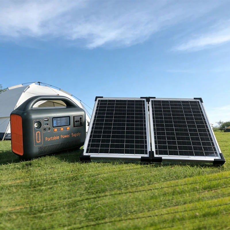 A dark gray portable power supply and a foldable solar panel set up on grass near a tent outdoors.