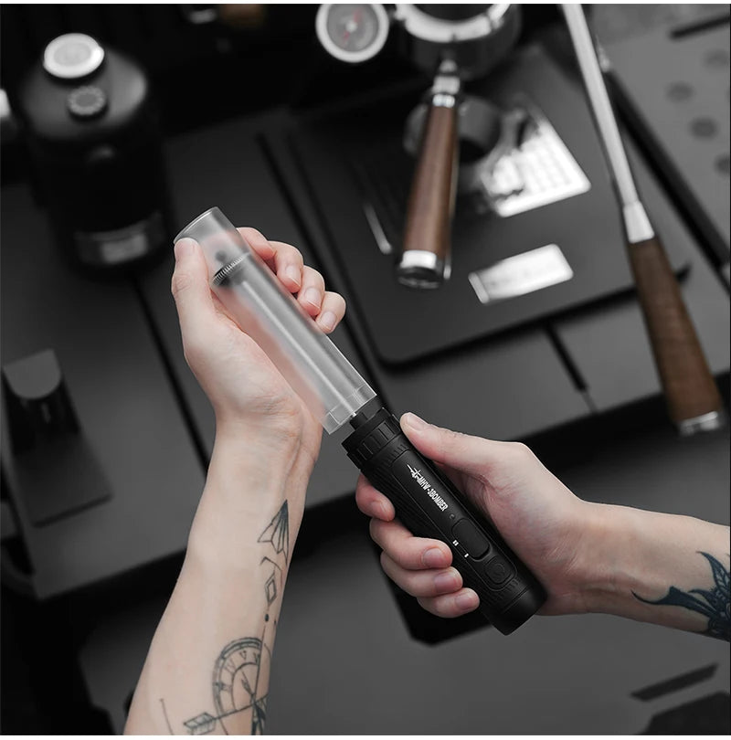 Hands holding an electric coffee grinder featuring a matte black handle and a clear, cylindrical top container.