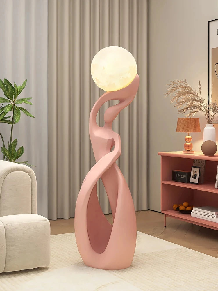 Abstract pink resin floor ornament featuring a stylised figure holding a glowing, moon-like orb lamp.