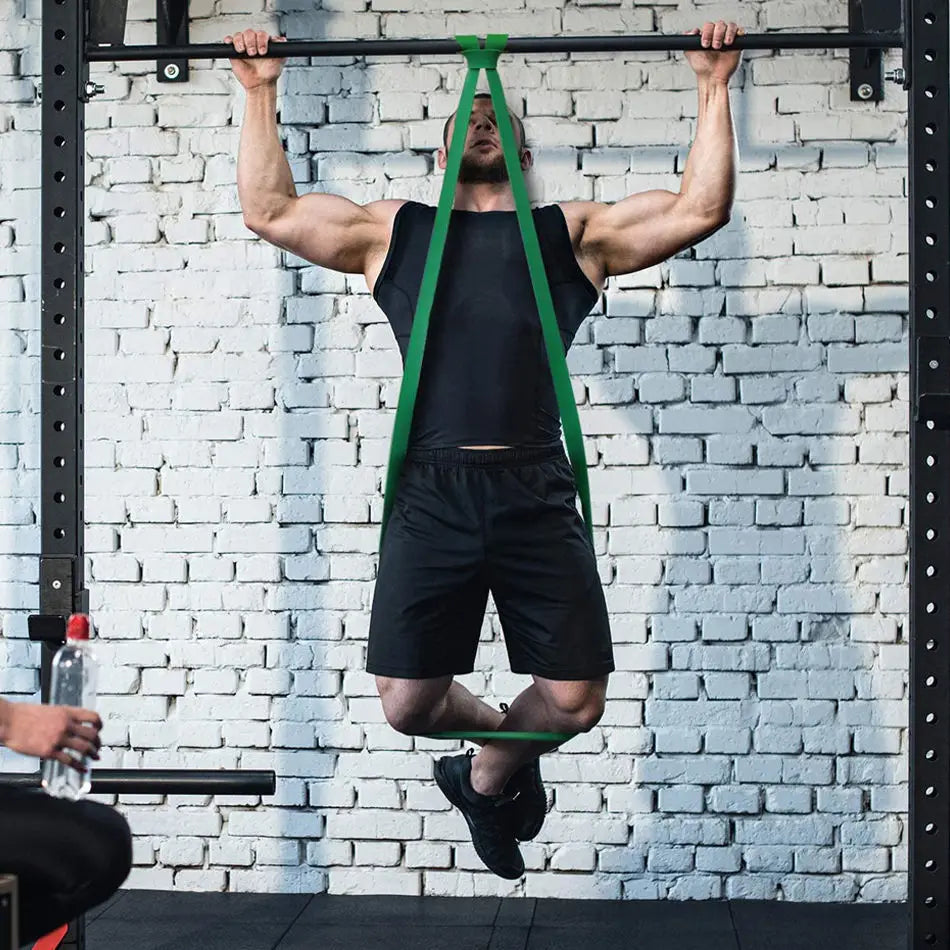 A man using a heavy-duty green resistance band looped over a pull-up bar for assisted pull-ups in a gym.