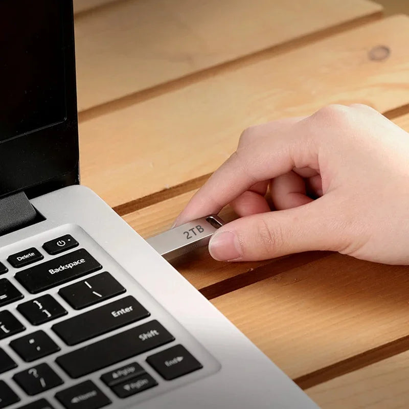A hand inserting a sleek, silver 2TB metal flash drive into a laptop port on a wooden desk.