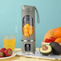 Portable sage-green electric blender bottle with a digital display, viewing window, and fruit inside, on a table.