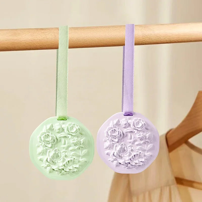 Two round wax sachets in light green and purple, featuring embossed floral details and matching hanging ribbons.