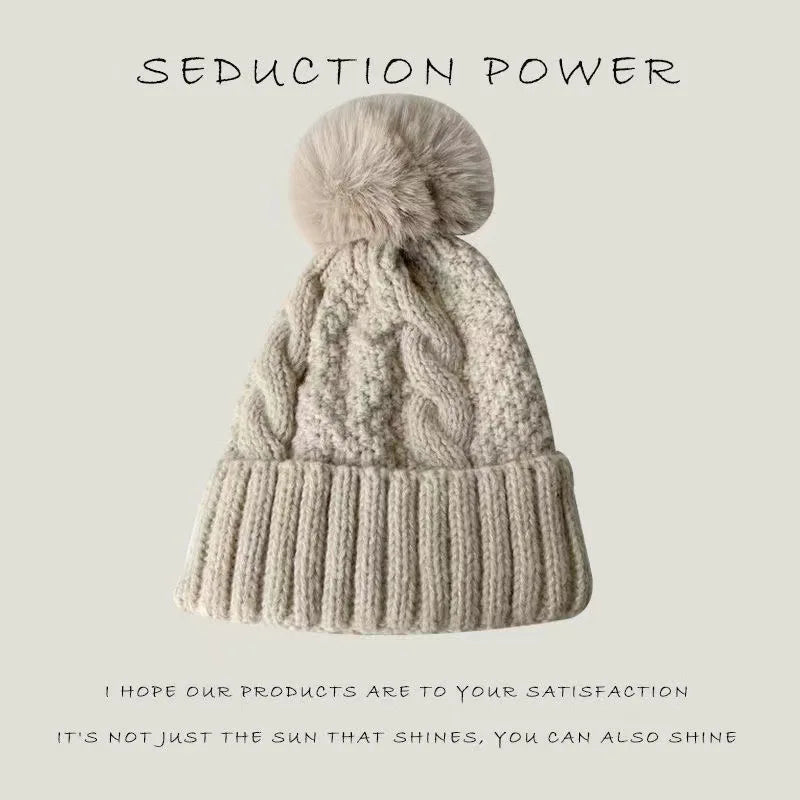 Beige cable-knit winter beanie hat with a fluffy faux fur pom-pom and a ribbed turn-up cuff.