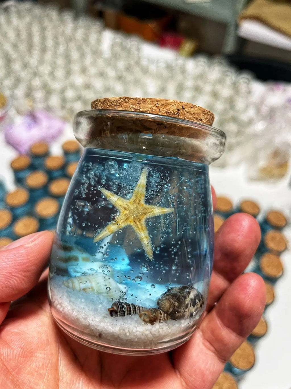 Small glass jar with cork lid, featuring a blue ocean-themed resin display with a starfish and seashells on sand.