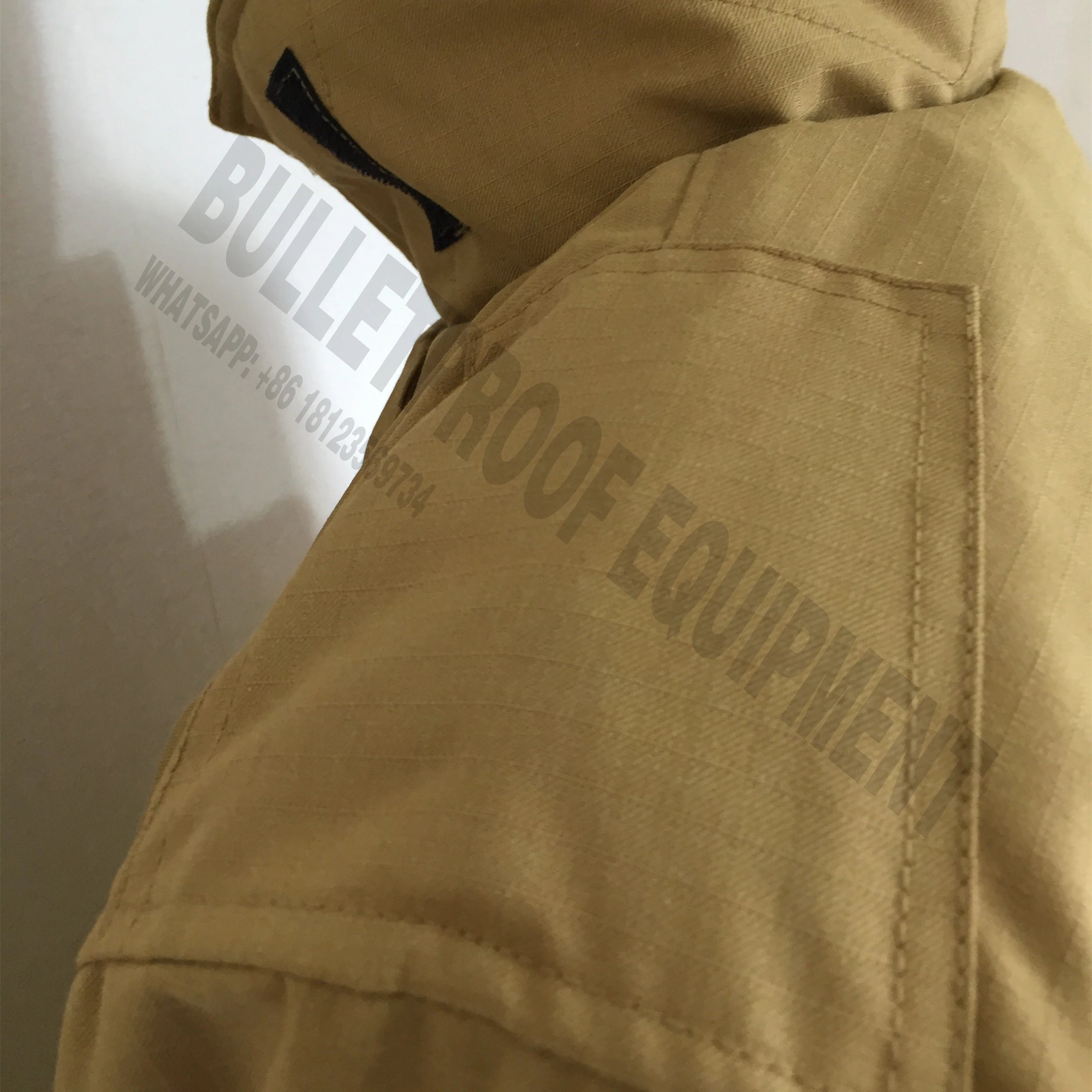 Close-up of a tan, ripstop fabric tactical jacket with a shoulder pocket and hood, against a light background.
