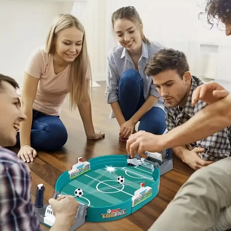 Friends playing a teal, oval-shaped tabletop football game with mini soccer balls on a wooden floor.