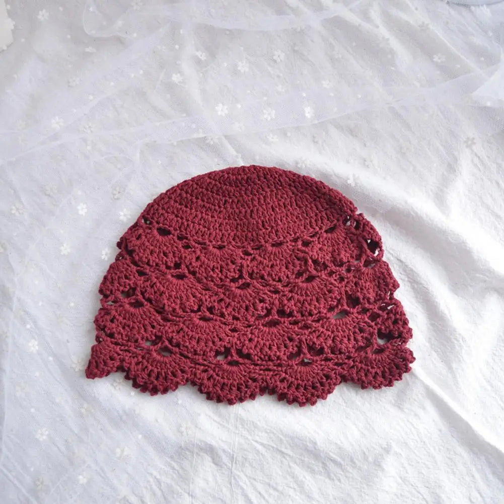 A deep burgundy crochet hat with an intricate hollow lace pattern and a scalloped edge, set on a white fabric backdrop.