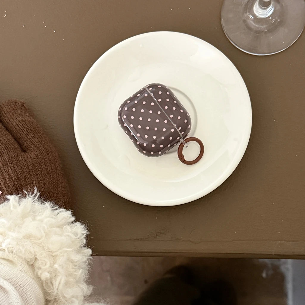 Brown, white polka-dot AirPods case with a circular ring attachment, resting on a white saucer.
