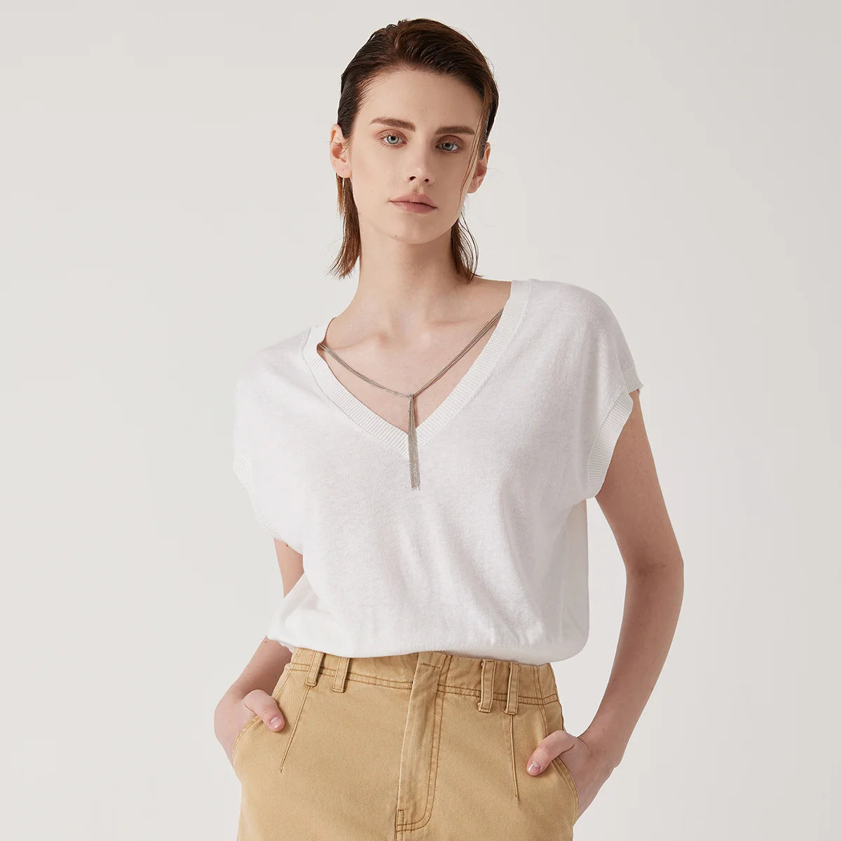 Model wearing a white V-neck knitted sleeveless top with a delicate silver chain detail, paired with khaki trousers.