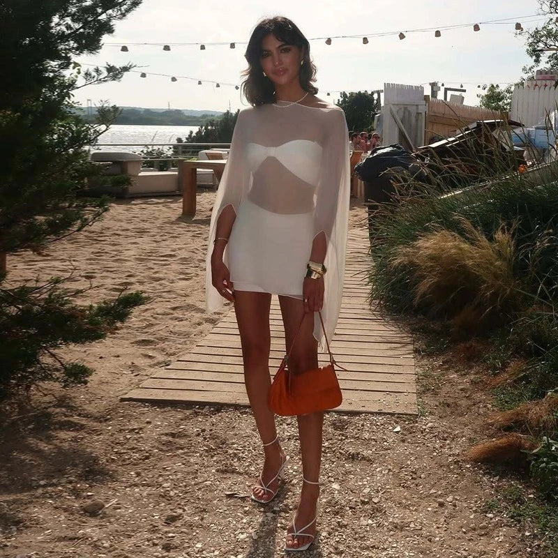 A white mini dress featuring a strapless bodice with a sheer, flowing cape-style overlay and matching strappy heels.