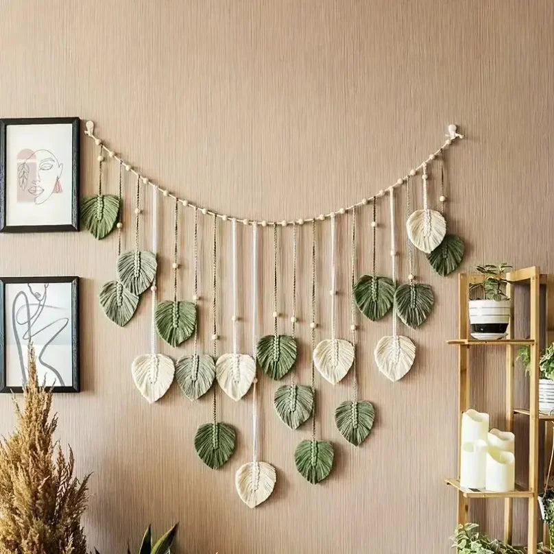 Boho macrame wall hanging featuring sage green and cream fabric leaves on a beaded string, styled in a living room.