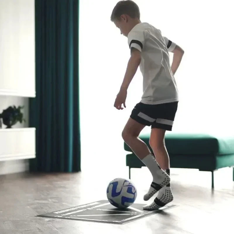 A boy in football gear practicing ball control on a black printed training mat indoors.