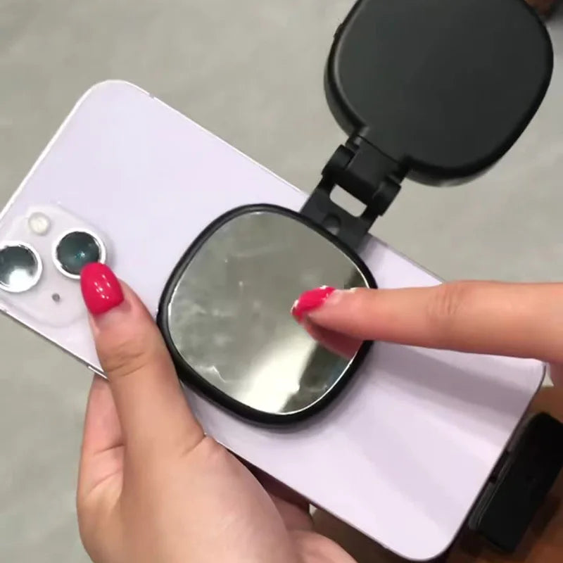 A hand holding a light purple smartphone with a black, foldable mirror attachment on the back.