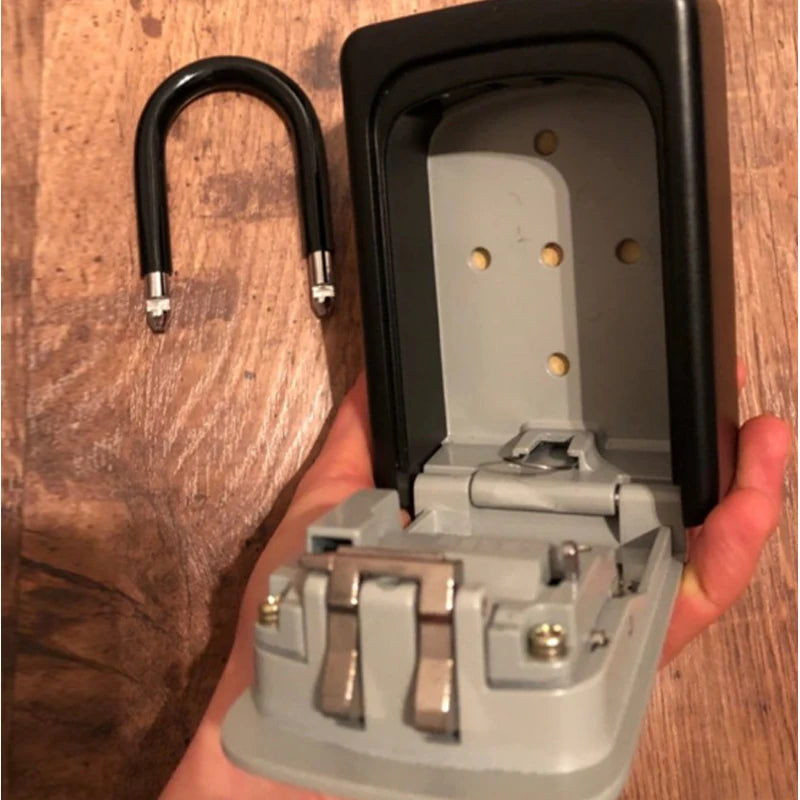 Black metal portable key lock box with a removable shackle, shown held in a hand against a wood background.