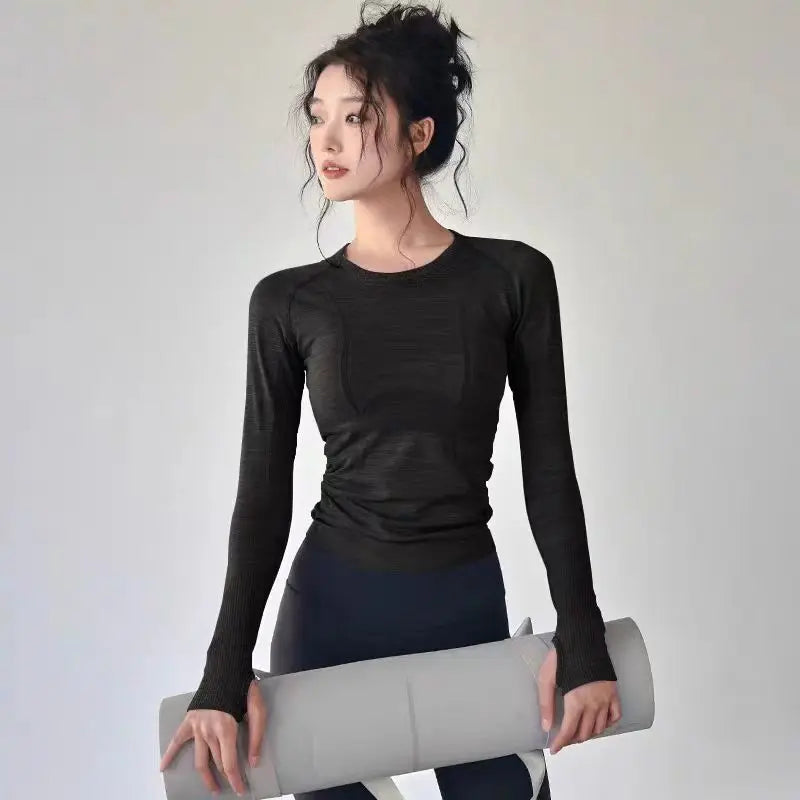 Woman wearing a black long-sleeve workout top with thumbholes, holding a grey yoga mat.