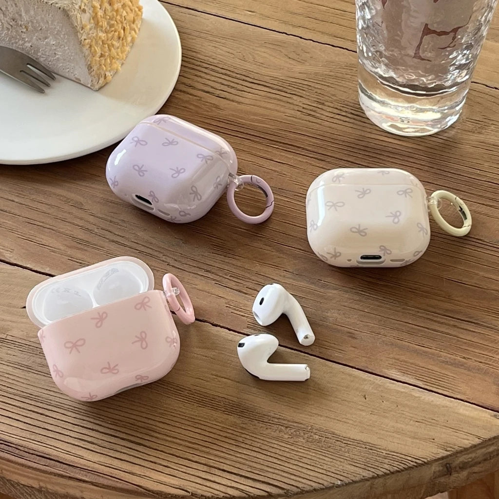 Glossy bow-patterned silicone AirPods cases in pastel purple, cream and pink with keyrings on a wooden table.