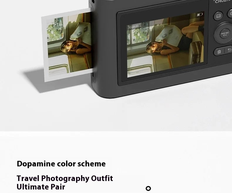 Matte dark grey digital instant camera ejecting a square photo print showing a person reading on a train.