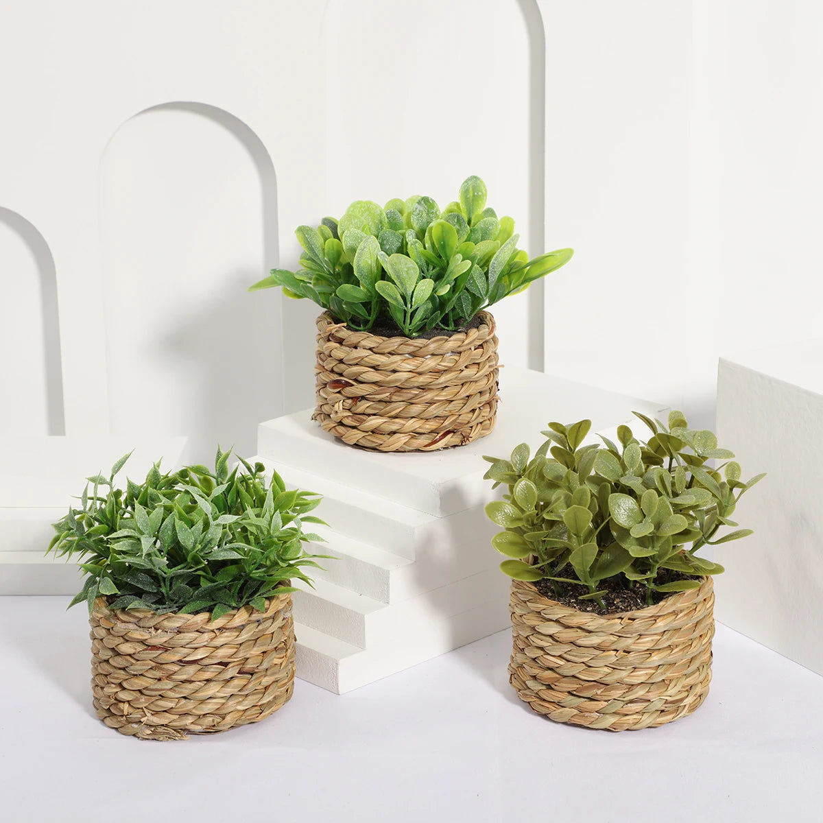 Three artificial green plants in small, woven seagrass pots arranged on a white tiered display.