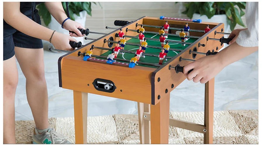 Two people playing a wooden-finish table football game with green pitch and colourful player figures.