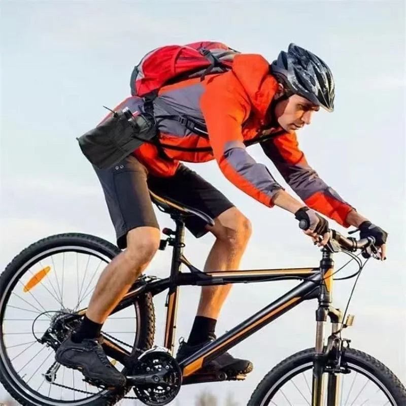 Cyclist wearing an orange and grey jacket with a red backpack, riding a black mountain bike against a clear sky.