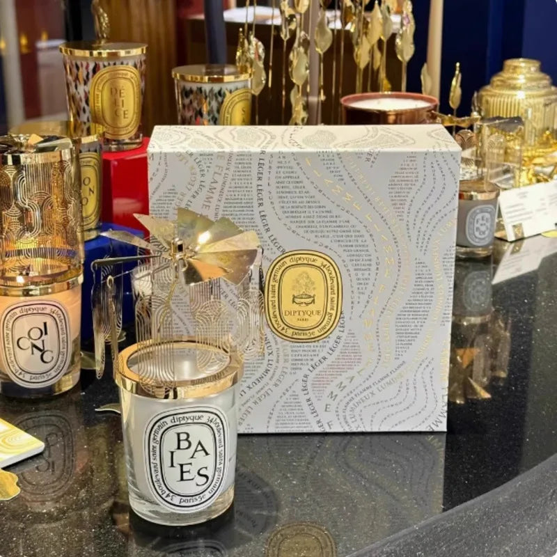 White scented candle in a glass jar with a gold-toned rotating metal carousel topper, beside a patterned gift box.