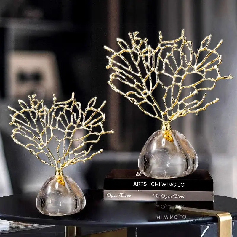 Two gold-branch coral sculptures on clear glass bases resting on a black table with books.
