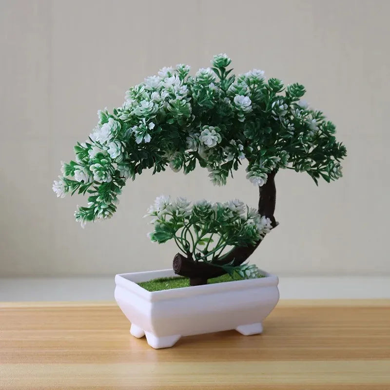 Artificial bonsai tree with green leaves and white flowers in a rectangular white pot on a wooden surface.