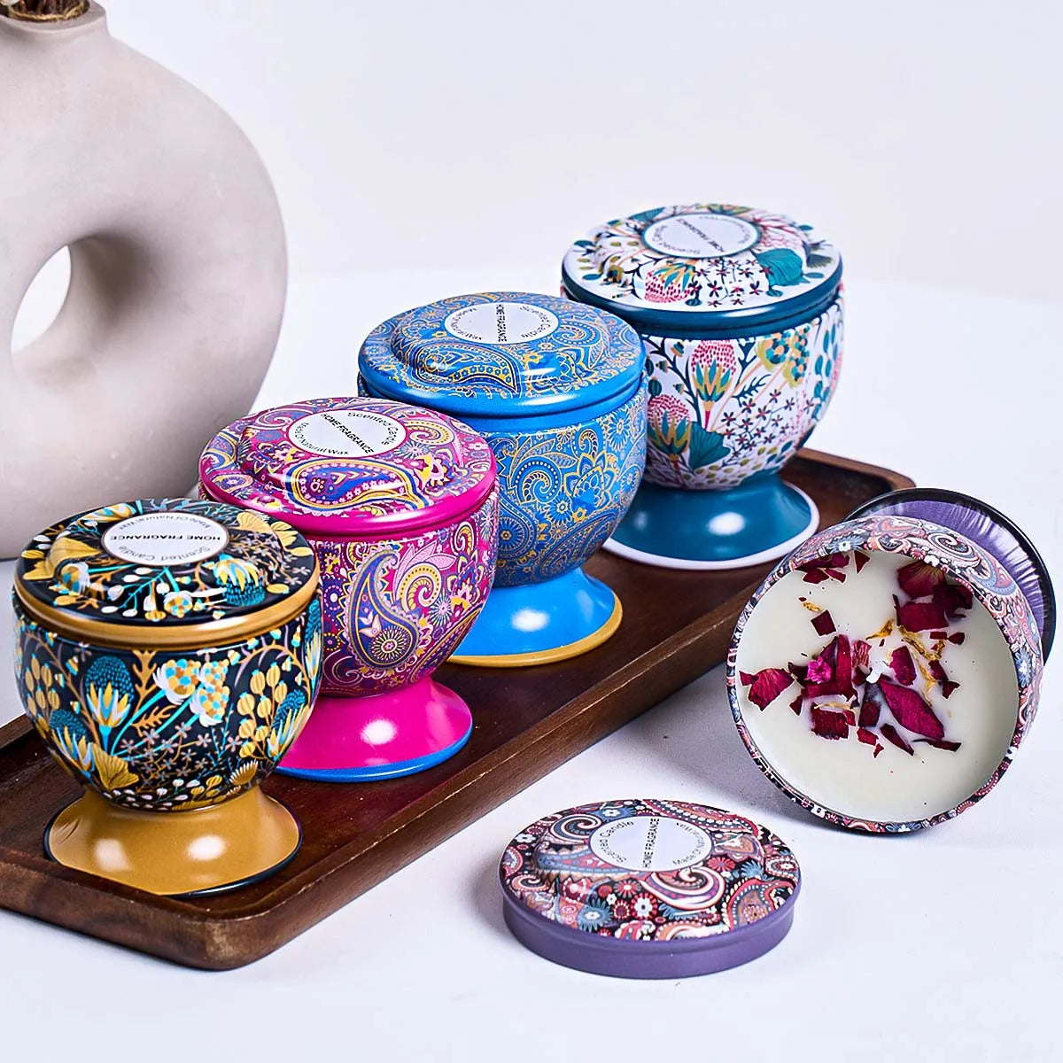 Set of four colourful, patterned metal candle tins with lids, featuring floral and paisley designs on a wooden tray.