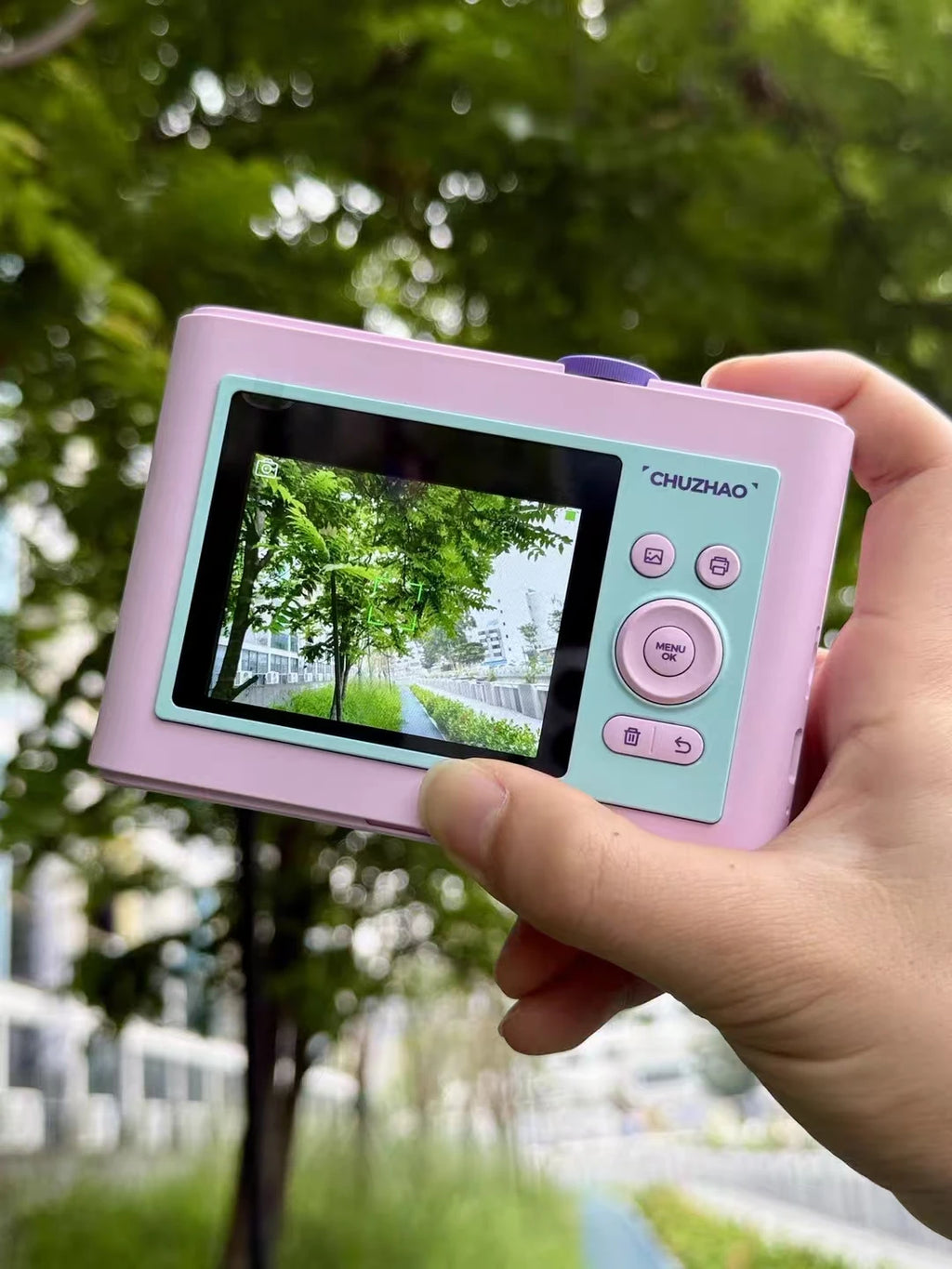 Pink and mint green plastic instant camera with a screen, physical buttons, and a purple top dial, held in a hand.