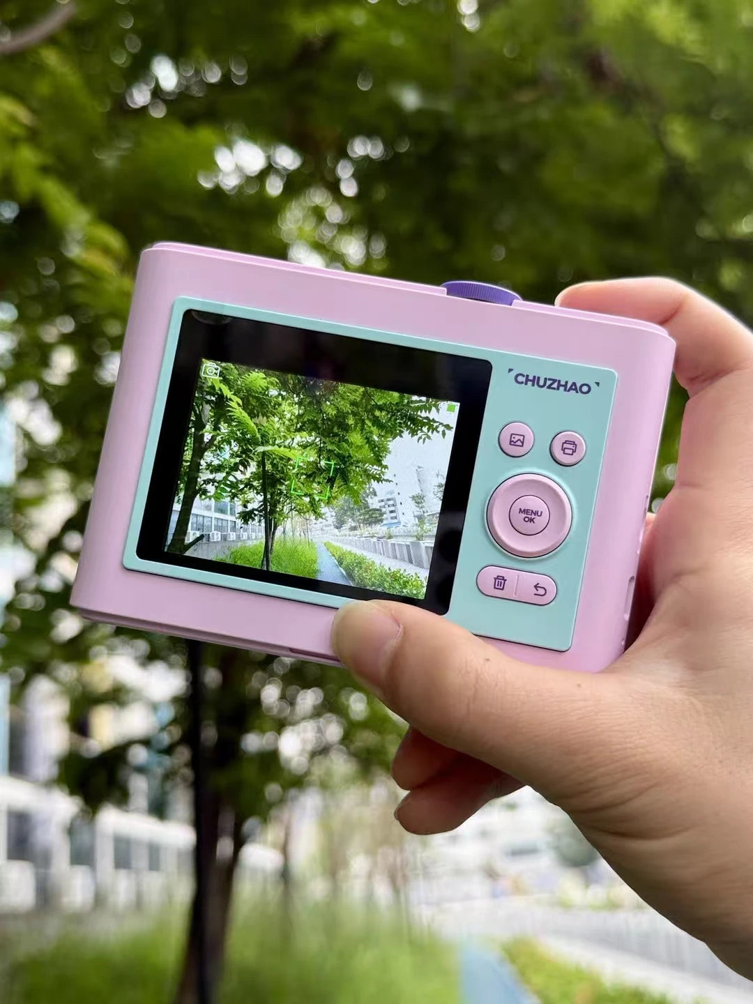 Pink and mint green plastic instant camera with a screen, physical buttons, and a purple top dial, held in a hand.