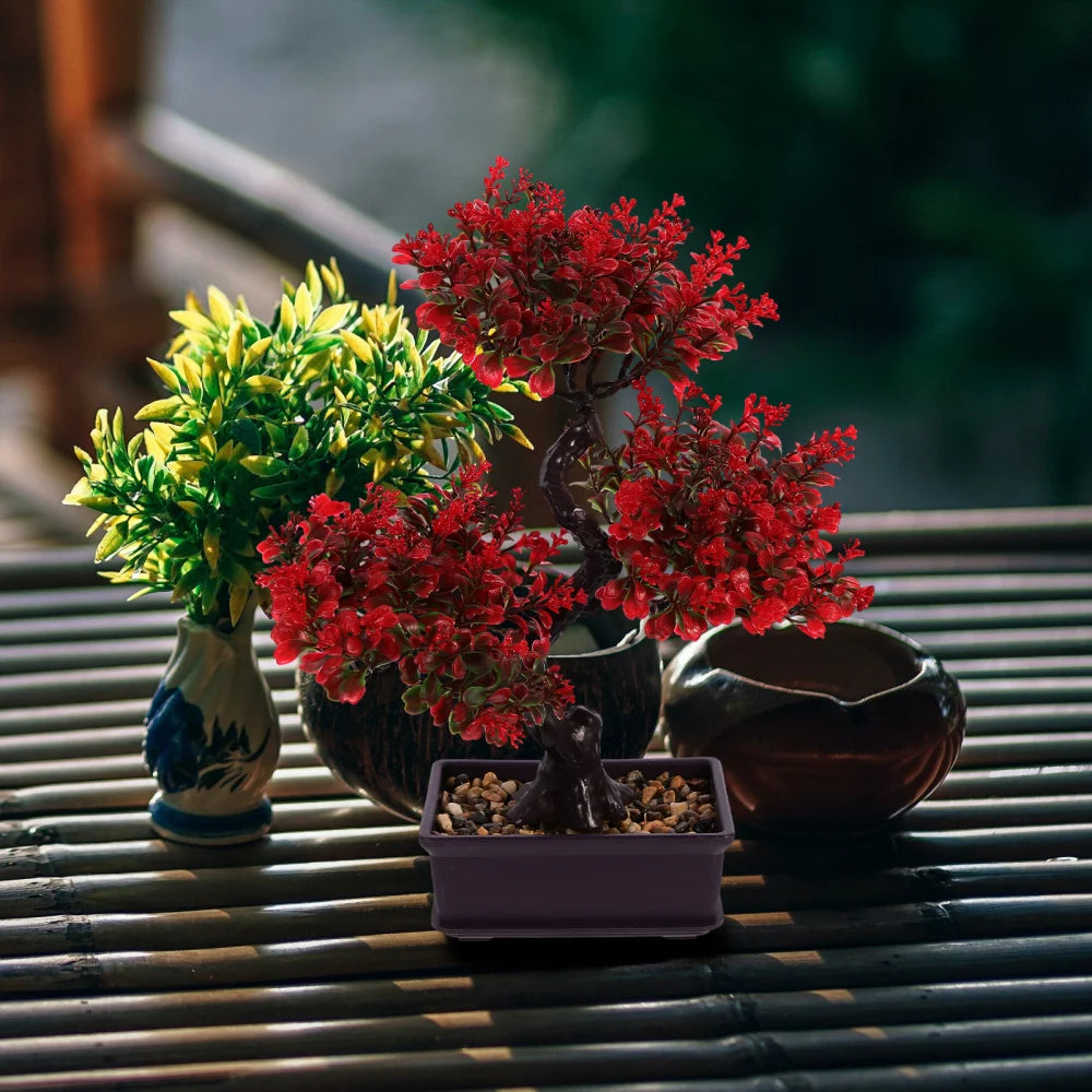 An artificial red bonsai tree in a rectangular pot sits on a slatted surface near a small yellow plant and bowls.